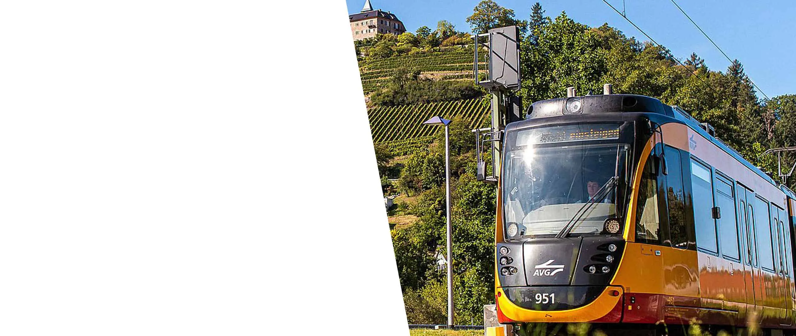 A yellow and red AVG light rail train passing a vineyard with a castle above