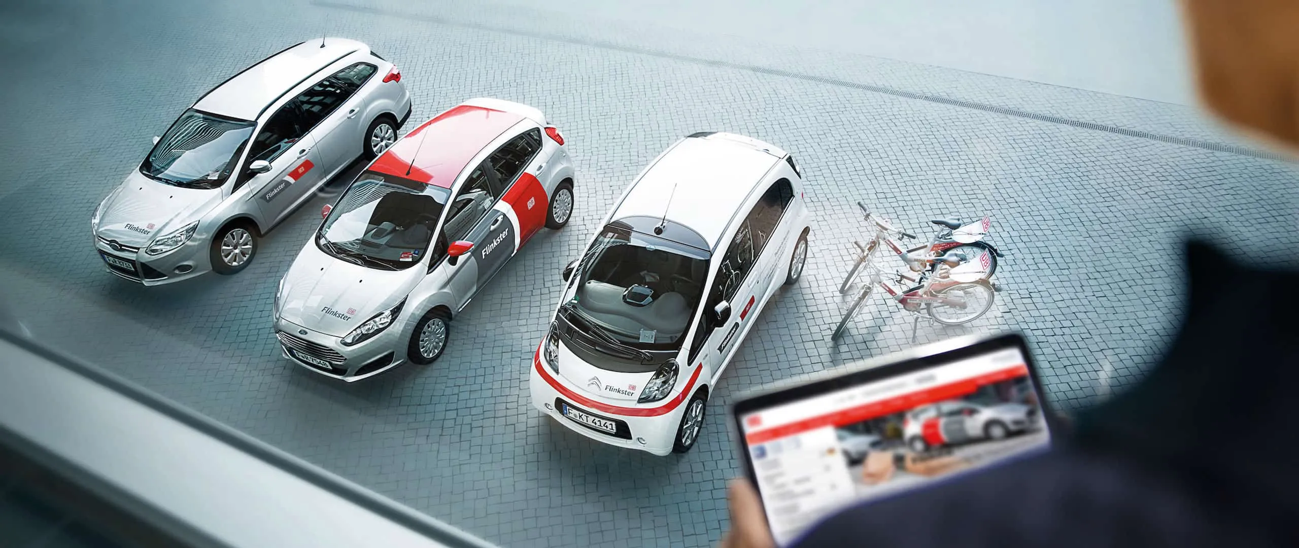 Drei Flinkster-Carsharing-Fahrzeuge und zwei Leihräder aus der Vogelperspektive, im Vordergrund ein Tablet mit der Buchungsansicht