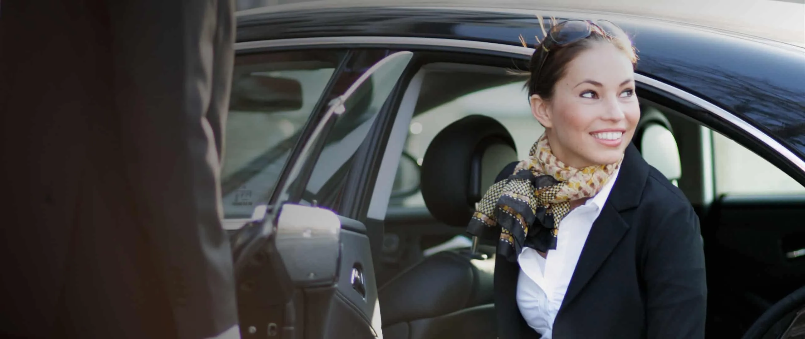 Fahrgast steigt lächelnd aus einer schwarzen Limousine, der Chauffeur hält die Tür