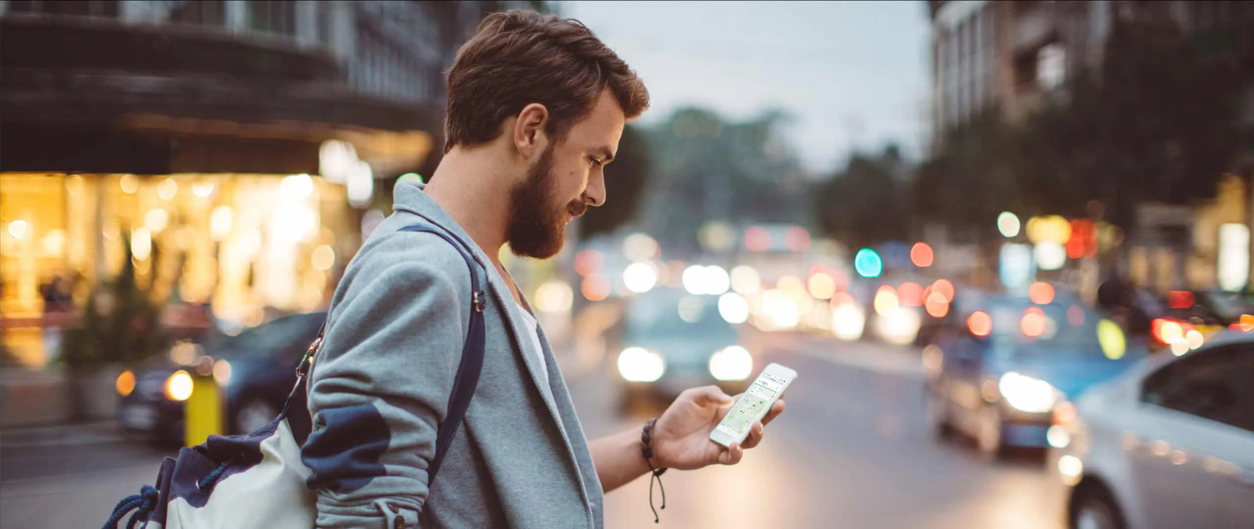 Mann mit Rucksack schaut abends an einer belebten Straße auf sein Smartphone