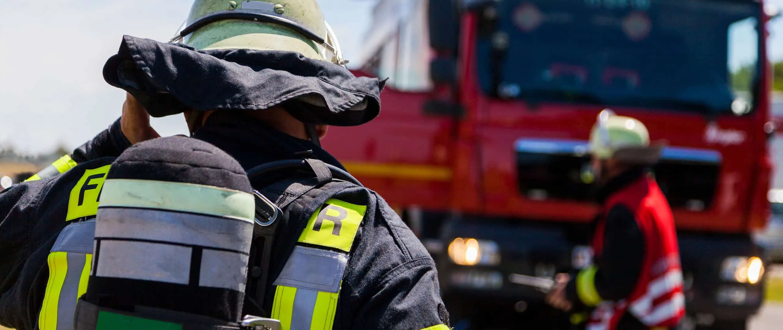Feuerwehrmann mit Atemschutzgerät von hinten, davor ein Löschfahrzeug im Einsatz