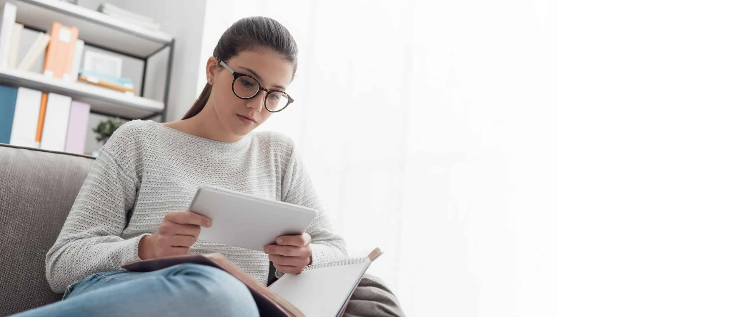 A young woman in glasses reads on a tablet, with an open book beside her